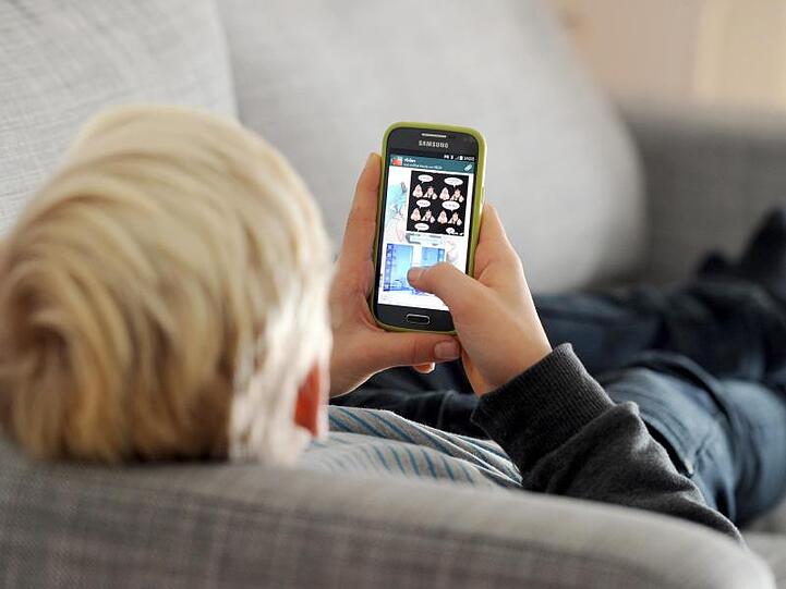 Ein Kind liegt auf einem Sofa und blickt auf sein Smartphone.