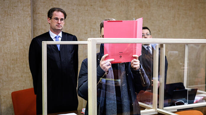 Der Angeklagte steht im Gerichtssaal vor seinem Rechtsanwalt Florian Schmidtke und verdeckt sein Gesicht.