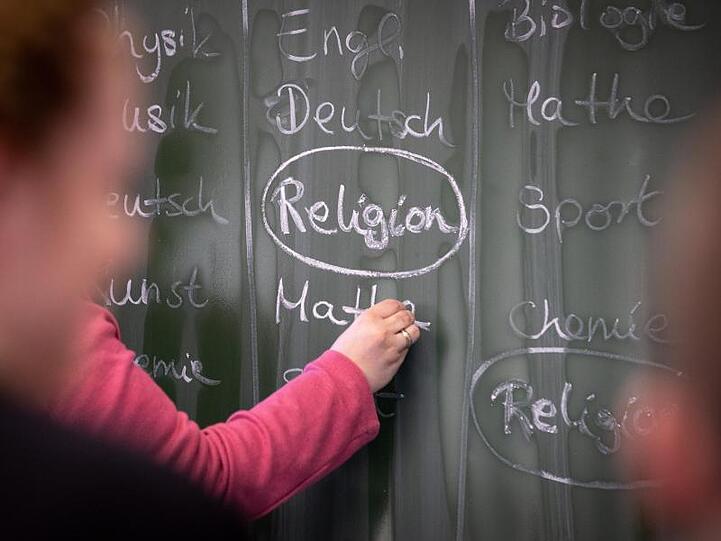 Eine Lehrerin schreibt mit Kreide einen Stundenplan mit Religionsunterricht an eine Tafel. Foto: Friso Gentsch/Archivbild