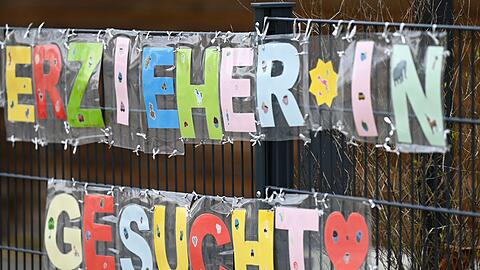 Mit bunten Buchstaben sind die Worte „Erzieher*in gesucht“ geformt, die an einem Zaun einer Kindertagesstätte befestigt sind.