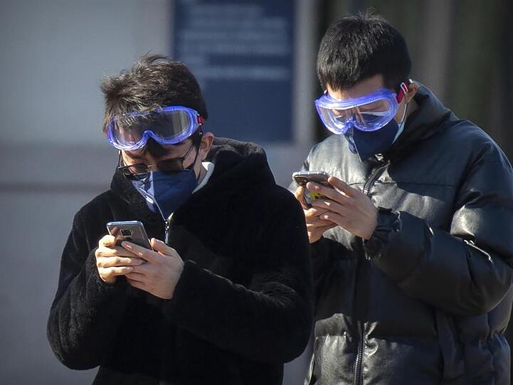 Reisende tragen Mundschutz und Schutzbrillen, während sie auf ihre Smartphones schauen. Foto: Mark Schiefelbein/AP/dpa/Archiv