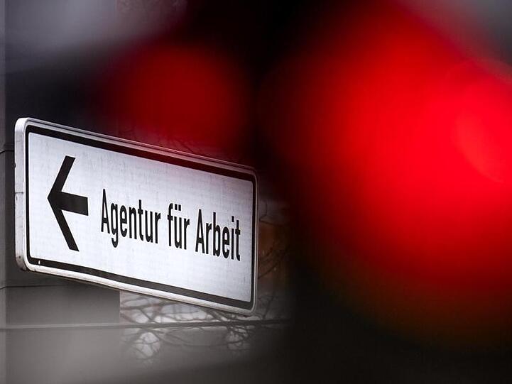 Eine rote Ampel leuchtet vor einem Schild, das auf die Agentur für Arbeit hinweist. Foto: Sina Schuldt/dpa/Symbolbild