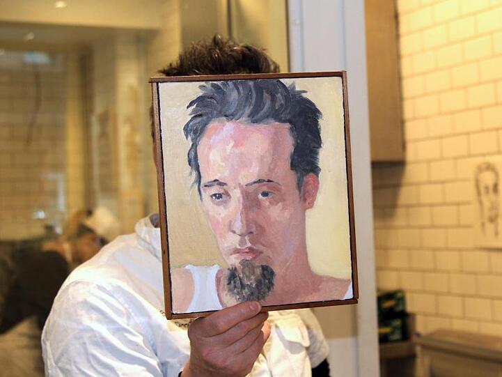 Sebastian Brecht hält sich ein Portrait vor sein Gesicht, das die Künstlerin Alix Bailey von ihm gemalt hat.