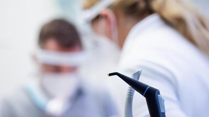 Ein Zahnarzt und Zahnarzthelferin mit Mund-Nasen-Schutz und Schutzvisieren versorgen eine Patientin.