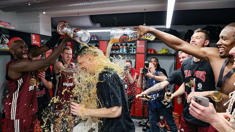 Der FC Bayern Basketball ist Pokalsieger 2024: Die Münchener Isaac Bonga (l) und Serge Ibaka (r) geben Andreas Obst (M) eine Bierdusche.
