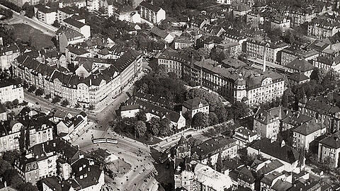Diese Aufnahme aus dem Jahr 1932 zeigt den Rotkreuzplatz mit Blickrichtung Nordwest. Der Krieg hat vonb der alten Bebauung nichts übriggelassen. In der Bildmitte ist das alte Neuhauser Jagdschlössl zu sehen, dahinter das Rotkreuzkrankenhaus. Das Türmchen links gehörte zur Winthirapotheke. Von unten kommt die Nymphenburger Straße ins Bild, sie zweigt nach rechts ab.
