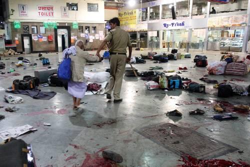 An der Hand führt ein Polizist einen alten Mann aus dem Chaos von Trümmern und Blut, das ein Terroranschlag auf den überfüllten Hauptbahnhof von Bombay hinterlassen hat.