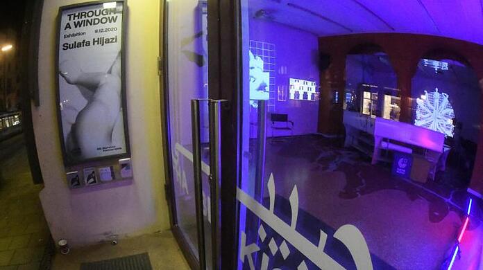 In blauem Licht leuchtet der Innenraum des Habibi Kiosk der Münchner Kammerspiele in der Maximilianstrasse, auf der eine Trambahn fährt.