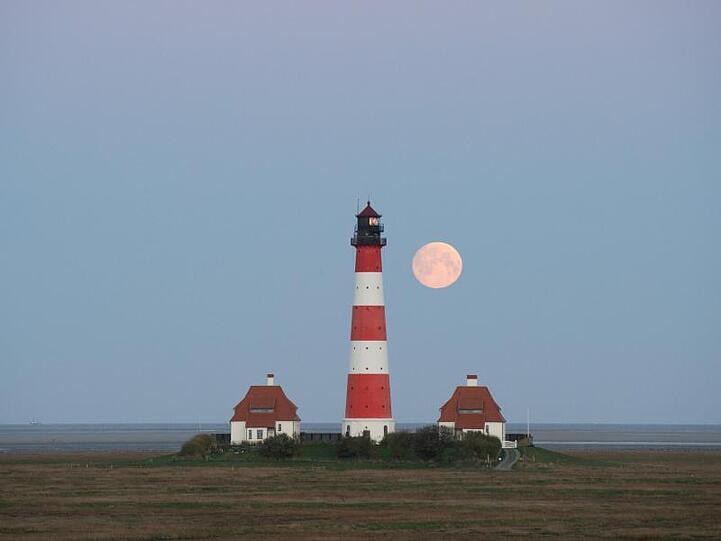 Der Mond neben dem Leuchtturm in Westerhever auf der nordfriesischen Halbinsel Eiderstedt.