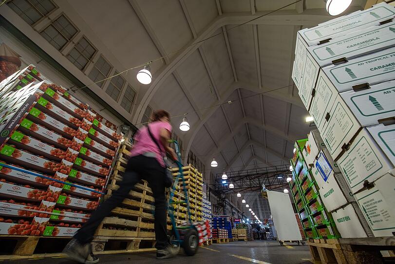 Eine Kundin geht mit einer Sackkarre in Halle 1 vom Großmarkt München an dem aufgestapelten Obst vorbei.