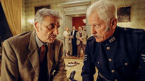 Chief Inspector Francis Lightmyer (Udo Wachtveitl, links vorne) und Constable Ivor Partridge (Miroslav Nemec, rechts vorne) untersuchen Arthur im Kaminzimmer. Lady Bantam (Sunnyi Melles), Charles (Ferdinand Hofer) und Dr. Mallard (Alexander Hörbe, rechts hinten) beobachten sie.