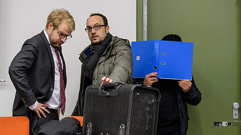 Prozessbeginn am Landgericht München: Ein Angeklagter versteckt sich hinter einem Aktenordner, während sich neben ihm Maximilian Donhauser (links), Rechtsanwalt einer mitangeklagten Frau, und Adam Ahmed, Rechtsanwalt des Hauptangeklagten, unterhalten.