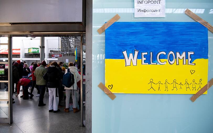 Ein Schild in den Farben der Ukraine mit der Aufschrift "Welcome" am Hauptbahnhof: im Hintergrund Helfer und Caritas-Mitarbeiter.