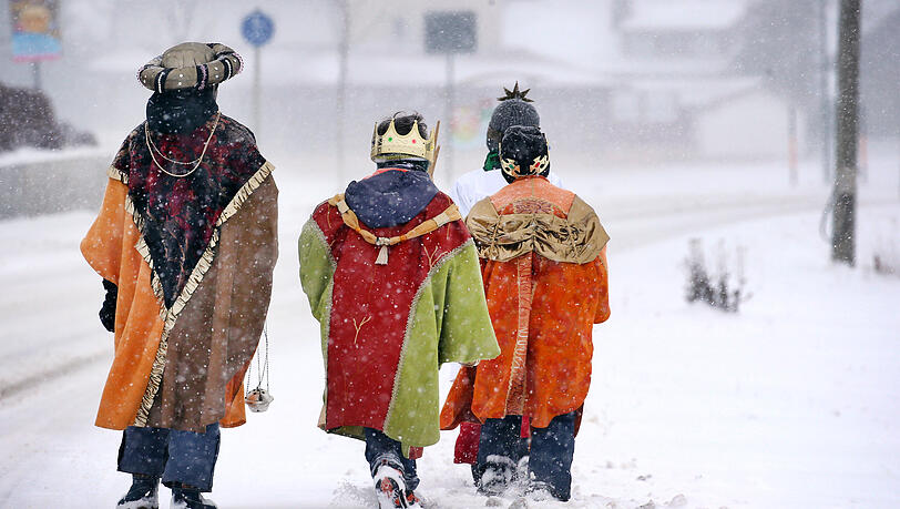 Sternsinger gehen im dichten Schneetreiben in ihren bunten Kostümen an einer Straße entlang. (Archivbild)