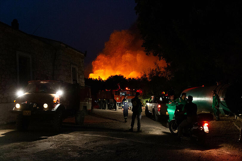 Athen: Einsatzkräfte stehen zur Lagebesprechung im Rahmen der Löscharbeiten während der Waldbrände an ihren Fahrzeugen. Im Hintergrund färben Flammen den Rauch, der aus einem Waldgebiet empor steigt.