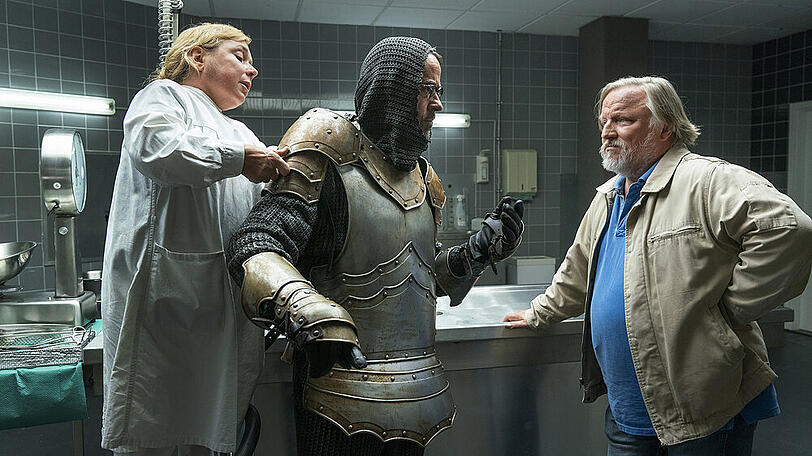 "Tatort: Es lebe der König!": Kommissar Frank Thiel (Axel Prahl, r.) hört, was Prof. Karl-Friedrich Boerne (Jan Josef Liefers) zu sagen hat, während ihm Silke Haller (ChrisTine Urspruch) in der Rechtsmedizin dabei hilft, die Ritterrüstung anzulegen.