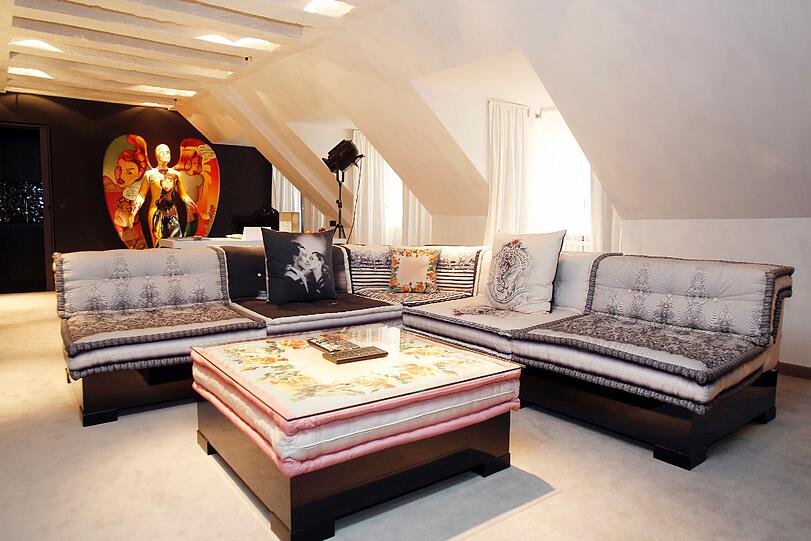 Die breite graue Couch ist von Jean-Paul Gaultier designt. Unter der Glasplatte des Tisches sorgt ein rosa Kissen für Farbkontraste.