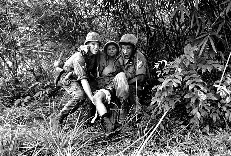 Um dem Feuer der Scharfschützen zu entkommen, tragen zwei amerikanische Ärzte einen verwundeten Fallschirmjäger zu einem Rettungshubschrauber (Juni 1965). Eine ganze Kompagnie war in einem Dschungel in der Nähe von Thoung Lang direkt in eine Sammelstelle der Vietcong geraten