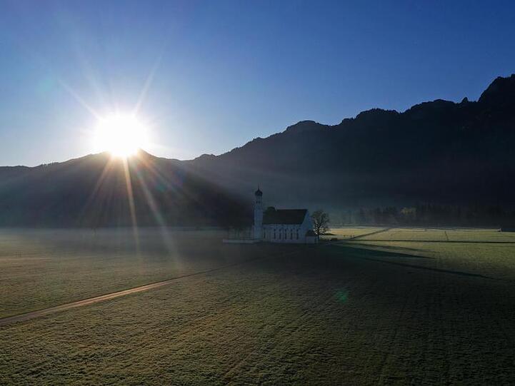 Die Sonne geht über den Bergen hinter der im leichten Morgennebel liegenden Wallfahrtskirche Sankt Coloman auf. Foto: Karl-Josef Hildenbrand/dpa/Archivbild