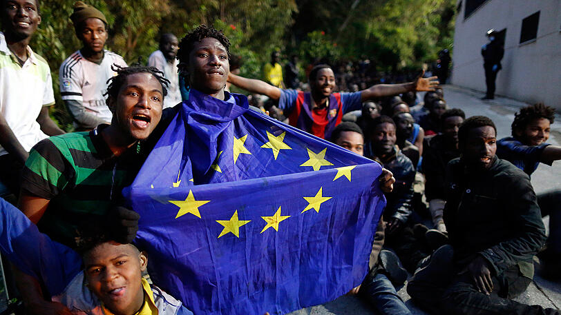 Flüchtlinge sitzen am 17.02.2017 in der spanischen Exklave Ceuta in Marokko mit einer EU-Flagge in den Händen auf dem Boden, nachdem sie am Morgen einen Grenzzaun gestürmt haben.