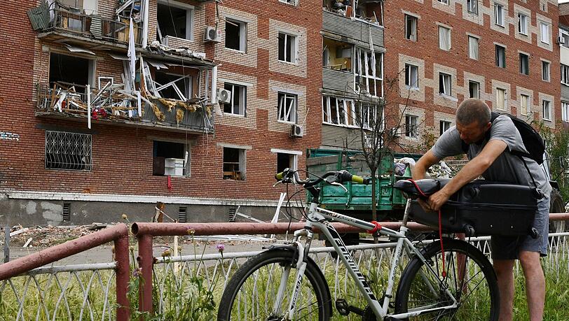 Ein Mann in Slowjansk zurrt einen Koffer auf seinem Fahrrad fest. Das Wohnhaus hinter ihm wurde bei einem nächtlichen Raketeneinschlag beschädigt.