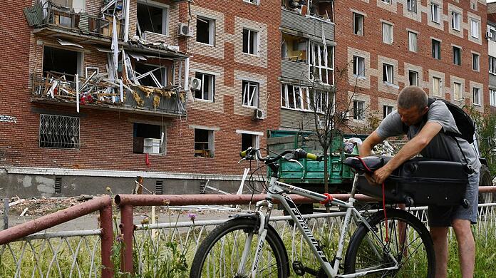 Ein Mann in Slowjansk zurrt einen Koffer auf seinem Fahrrad fest. Das Wohnhaus hinter ihm wurde bei einem nächtlichen Raketeneinschlag beschädigt.
