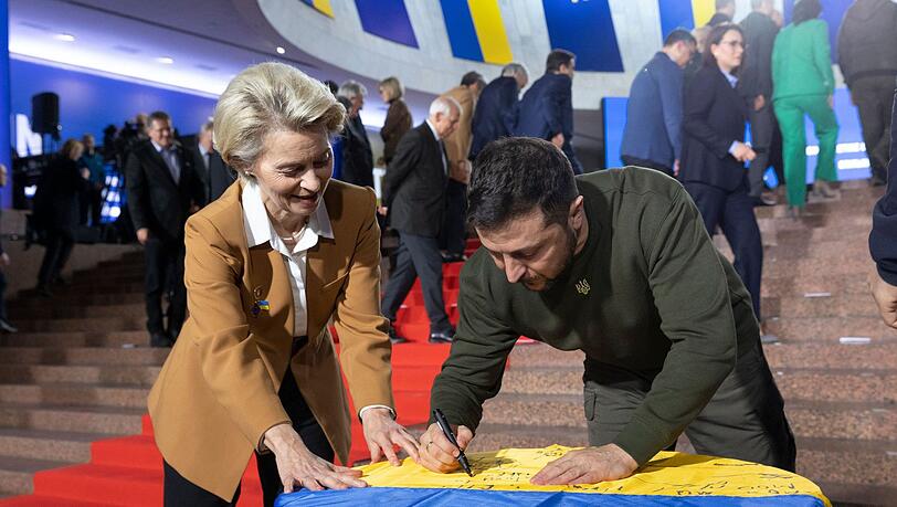 Wolodymyr Selenskyj und Ursula von der Leyen schreiben ihre Wünsche auf eine ukrainische Flagge.