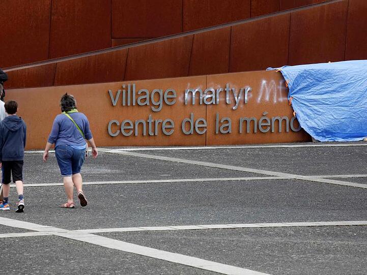 Besucher gehen an einem abgedeckten Graffiti auf dem Schriftzug am Eingang der Gedenkstätte in Oradour-sur-Glane vorbei. Unbekannte hatten unter anderem das Wort "Märtyrer" im Schriftzug mit weißer Farbe durchgestrichen und "Lügner" daneben geschrieben.