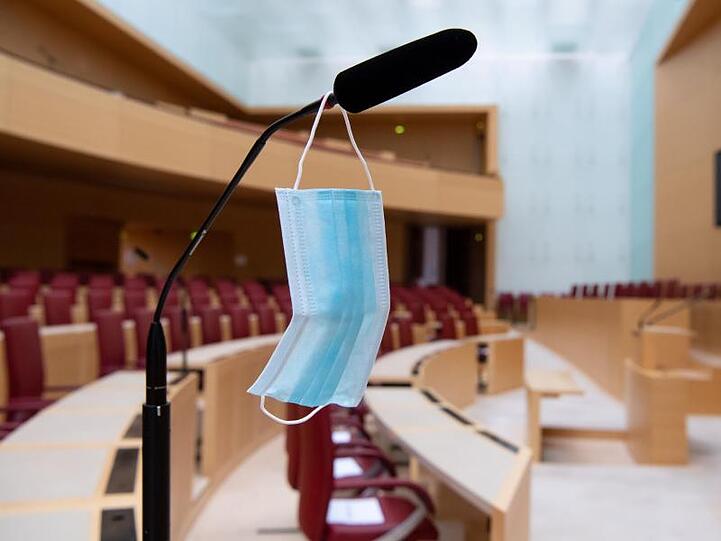 Ein Mund-Nase-Schutz hängt im Plenarsaal im bayerischen Landtag an einem Mikrofon. Foto: Sven Hoppe/dpa/Archiv/Illustration