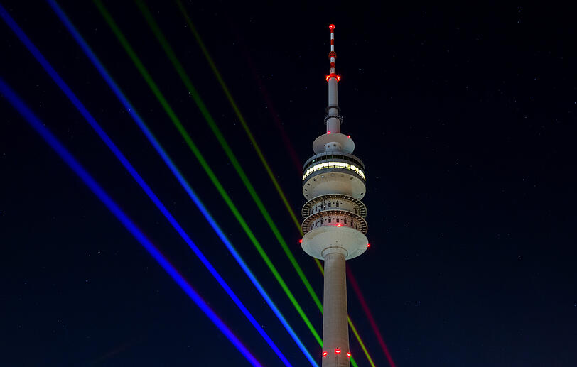 Laserstrahler projizieren bunte Strahlen neben dem Olympiaturm in den Nachthimmel. Sie sollen die Regenbogenfarben symbolisieren.