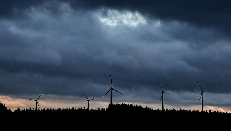 Windräder stehen unter dichten Regenwolken hinter einem Wald.