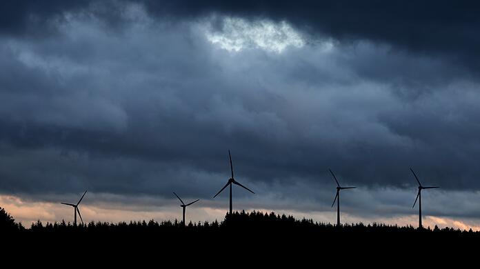 Windräder stehen unter dichten Regenwolken hinter einem Wald.