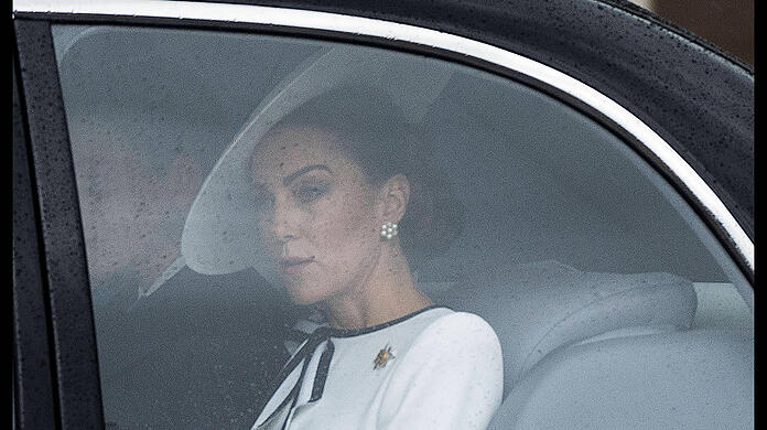 Der Samstagmorgen in London ist regnerisch. Kate ist gutbehütet. Sie ist in Weiß gekleidet. Ihr Outfit hat eine schwarz-weiße Schleife am Kragen.