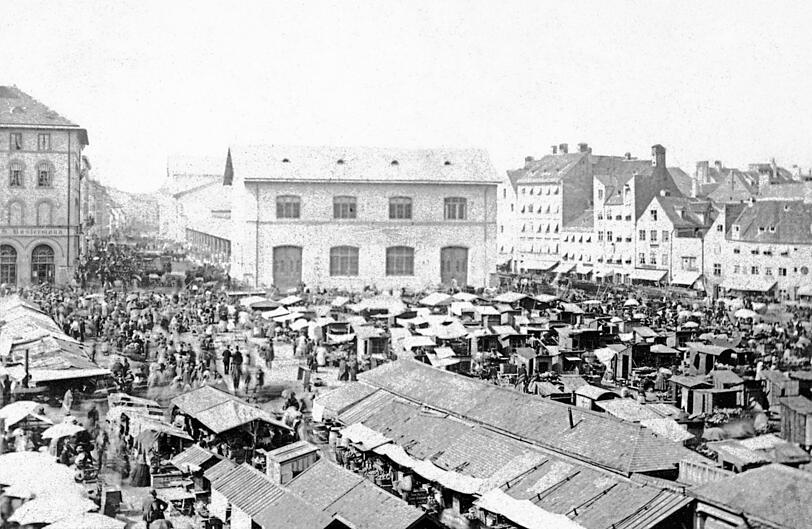 Ganz sch&ouml;n was los auf dem Viktualienmarkt 1865: viele kleine Standl, dahinter die Schrannenhalle und ganz rechts noch viel von der alten mittelalterlichen Bebauung.
