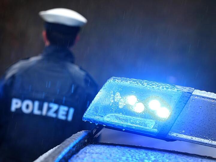 Ein Polizist steht im Regen vor einem Streifenwagen dessen Blaulicht aktiviert ist.