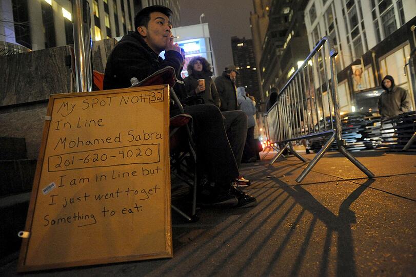 Auf dem Schild dieses rauchenden Herrn steht: "Mein Platz Nr. 32 - In der Schlange: Mohamed Sabra - 201-620-4020 - Ich stehe Schlange aber gerade bin ich etwas essen gegangen."