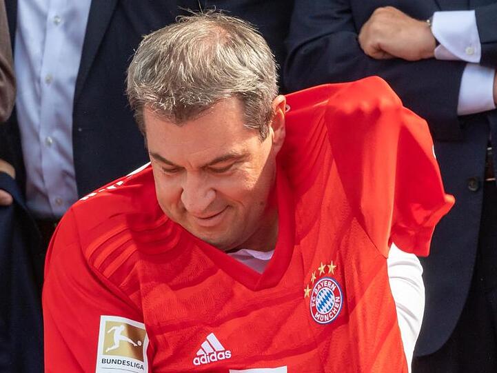 Bayerns Ministerpräsident Markus Söder zwängt sich in ein Trikot des FC Bayern München, das ihm zuvor Uli Hoeneß überreicht hat.
