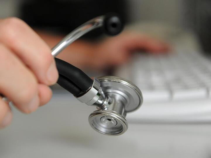 Ein Arzt hält ein Stethoskop in der Hand; mit der anderen Hand bedient er eine Computertastatur. Foto: Marijan Murat/Archivbild