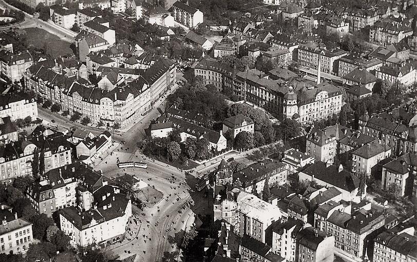 Die Aufnahme aus dem Jahr 1932 zeigt den Rotkreuzplatz mit Blickrichtung Nordwest. In der Bildmitte ist das alte Neuhauser Jagdschlössl zu sehen, dahinter das Rotkreuzkrankenhaus. Das Türmchen links gehörte zur Winthirapotheke. Von unten kommt die Nymphenburger Straße ins Bild, sie zweigt nach rechts ab.