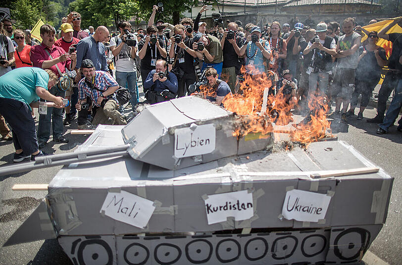 Fotografen, Reporter und Kameraleute umringen einen brennenden Panzer aus Pappe, auf dem die Ländernamen der Krisenherde Lybien, Mali, Kurdistan und Ukraine stehen, während einer Demonstration vor dem Marshall Center for European Security Studies in Garmisch-Partenkirchen.