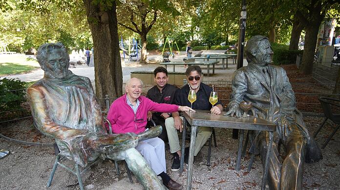 Noch ist Helmut Dietl am Café Münchner Freiheit neben dem Monaco Franze verhüllt. Die AZ durfte die Skulptur bereits unverhüllt fotografieren. Im Bild (v.l.): Kaffeehausgründer Karl Eisenrieder (83) mit Sohn Max Eisenrieder sowie Künstler Nikolai Tregor, der beide Skulpturen erschaffen hat.