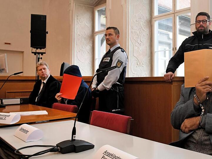 Der Vater (r) auf der Anklagebank mit Aktendeckel vor dem Gesicht, links einer der angeklagten Söhne. Foto: Stefan Puchner/dpa/Archivbild