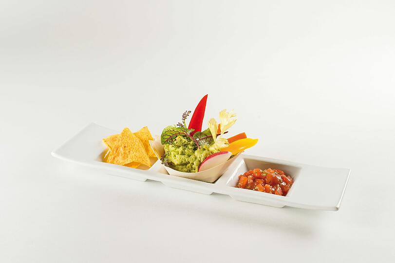 Das Menü zu Eloize - Vorspeise: Pikante Guacamole &ndash; im Holzschiffchen serviert &ndash; gespickt mit Gemüsesticks, dazu mit frischem Koriander fein abgeschmecktes Tomaten-Crudo und Taco-Chips