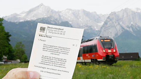Ein Mann hält ein 9-Euro-Ticket in der Hand, im Hintergrund fährt vor der Bergkulisse des Wettersteingebirges ein Zug vorbei.
