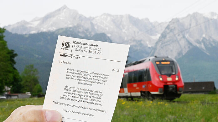 Ein Mann hält ein 9-Euro-Ticket in der Hand, im Hintergrund fährt vor der Bergkulisse des Wettersteingebirges ein Zug vorbei.