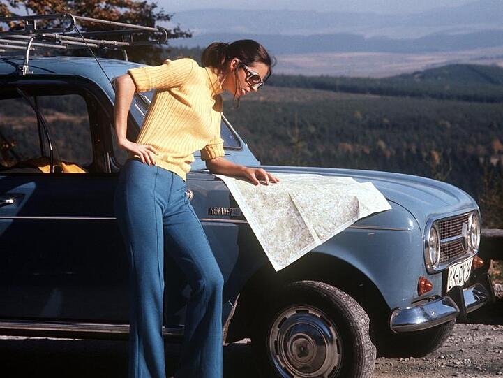 Eine junge Frau steht vor ihrem Wagen und informiert sich anhand einer Straßenkarte (Aufnahme aus den 70er Jahren).s").