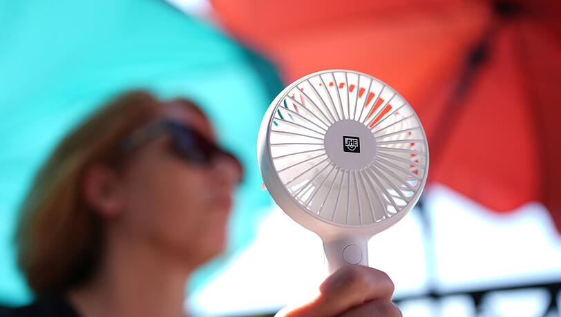 Eine Frau k&uuml;hlt sich mit einem Handventilator unter Sonnenschirmen auf einem Balkon in der Mittagssonne ab. Die Hitzewelle in Deutschland sorgt f&uuml;r schwei&szlig;treibende Temperaturen.