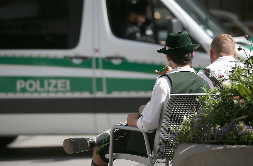 Zwei Männer in Tracht sitzen vor einem Polizeifahrzeug in der Münchner Fußgängerzone.