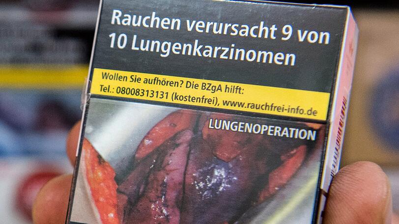 Ein Mann hält eine Zigarettenschachtel auf der ein Schockfoto einer Lungenoperation und der Warnhinweis "Rauchen verursacht 9 von 10 Lungenkarzinomen" aufgedruckt sind.