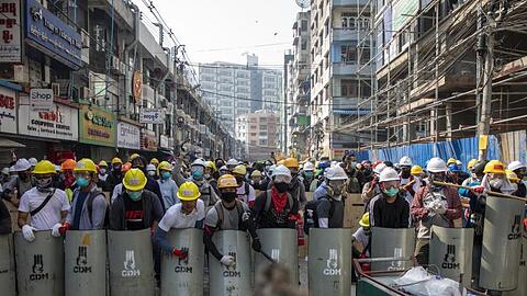 Mit Helmen und Schutzbrillen ausgerüstet schlagen die Demonstranten auf handgefertigte Schilder und bilden eine Schutzwand gegen die Polizei.